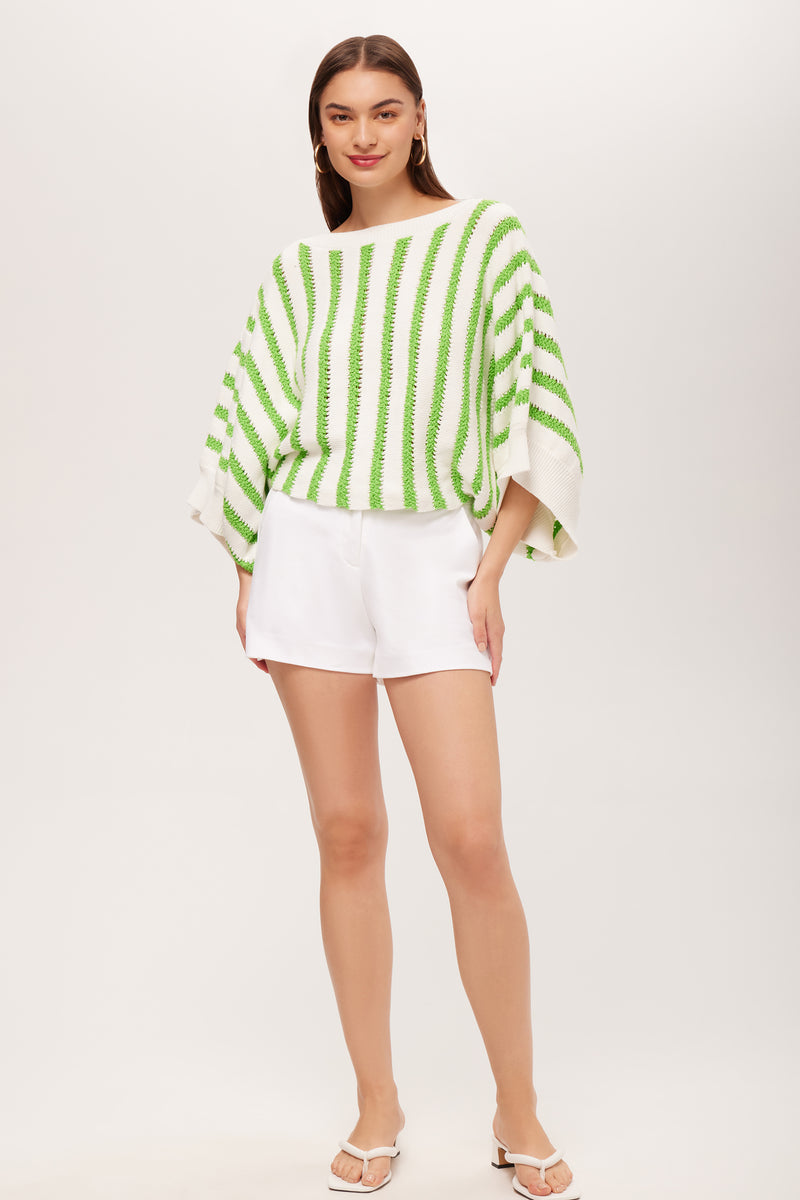 A woman with straight brown hair smiles slightly, wearing the Horizon 3/4-Sleeve Striped Boatneck Pullover—a loose white sweater with green stripes—paired with white shorts and sandals, standing against a plain background with one hand in her pocket.