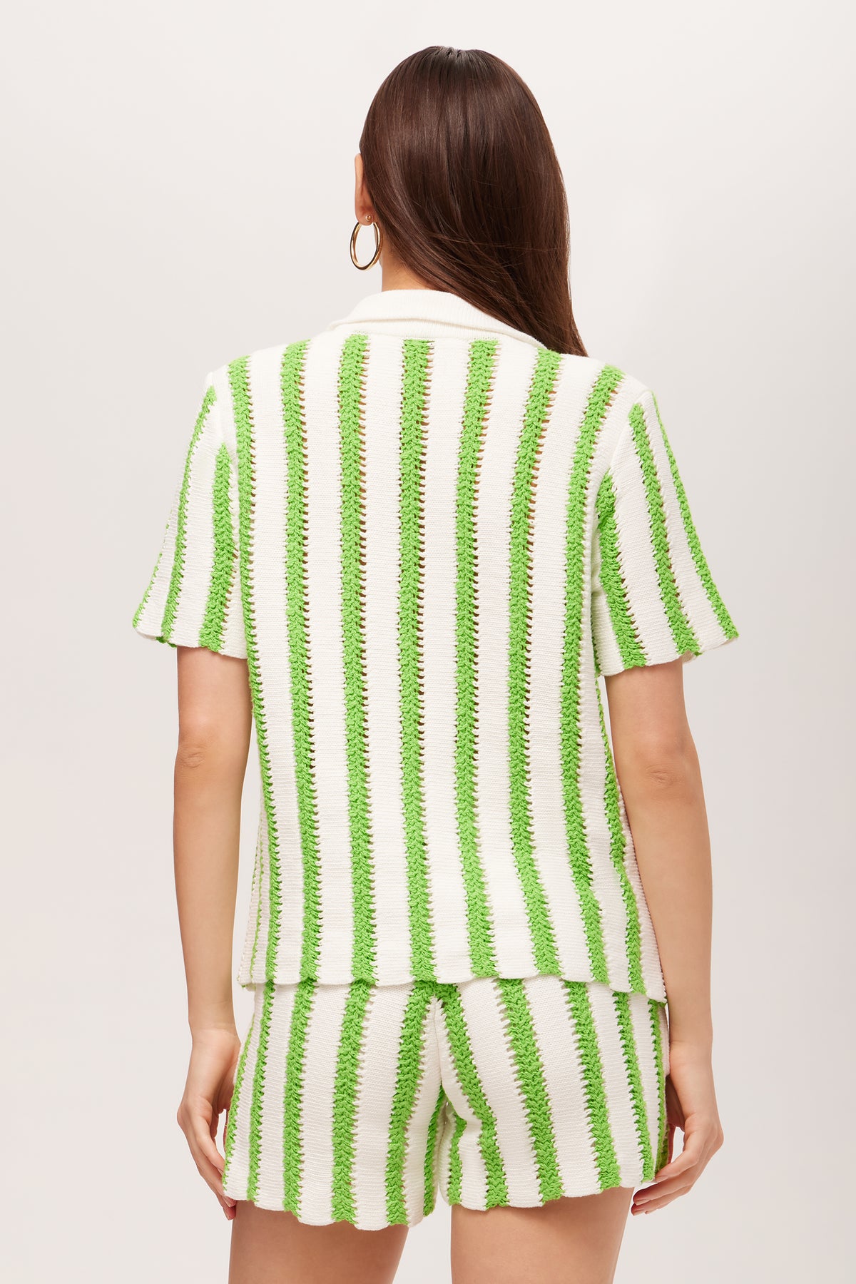 A woman with long brown hair and hoop earrings models the Sloane Short-Sleeve Striped Button-Up Shirt, a white top with vertical green stripes, shown from the back against a plain background.