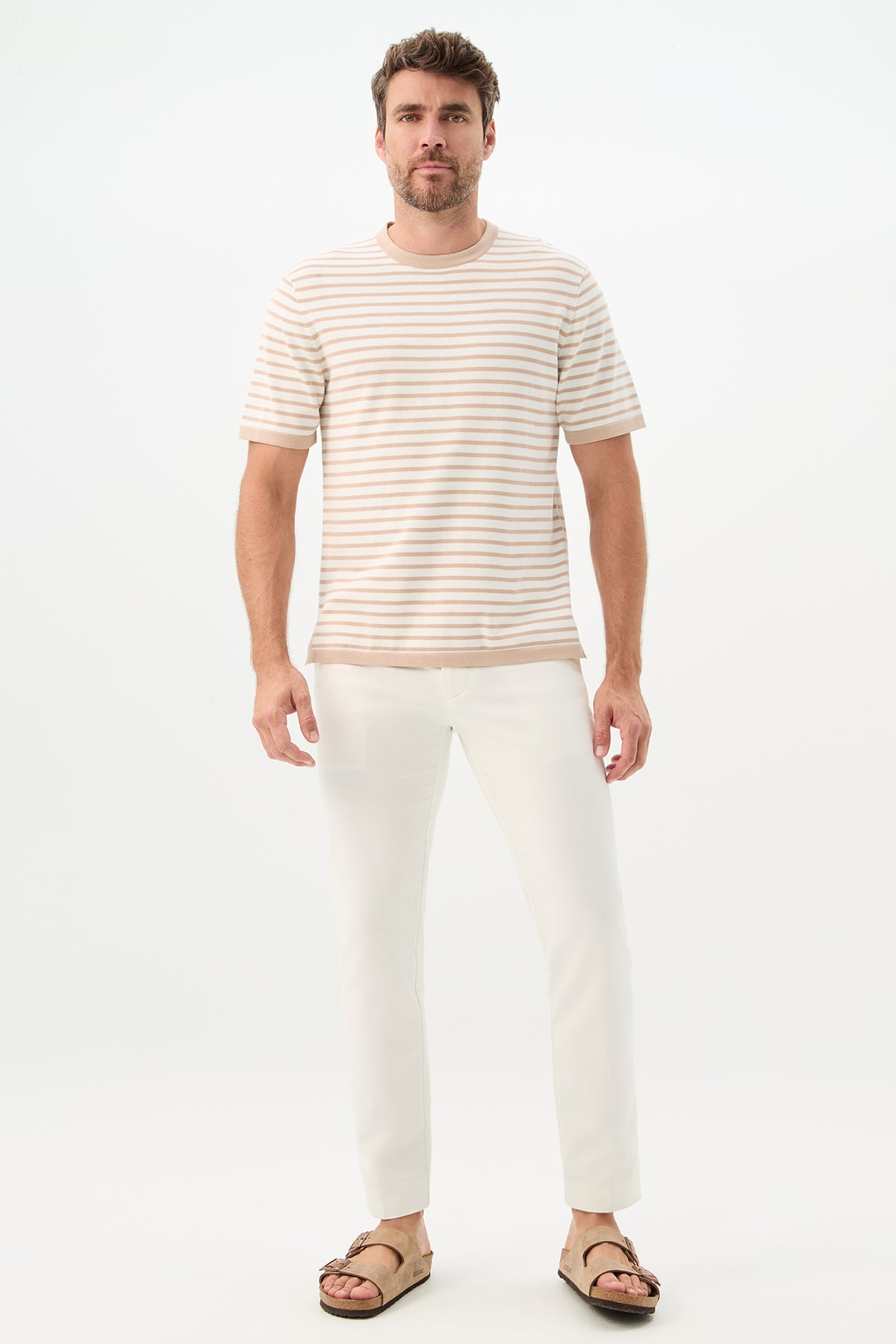A man with short hair and a trimmed beard stands against a plain white background, wearing the Giovanni Stripe Short-Sleeve Pullover in beige and white stripes, paired with white pants and brown sandals, looking straight ahead with a neutral expression.