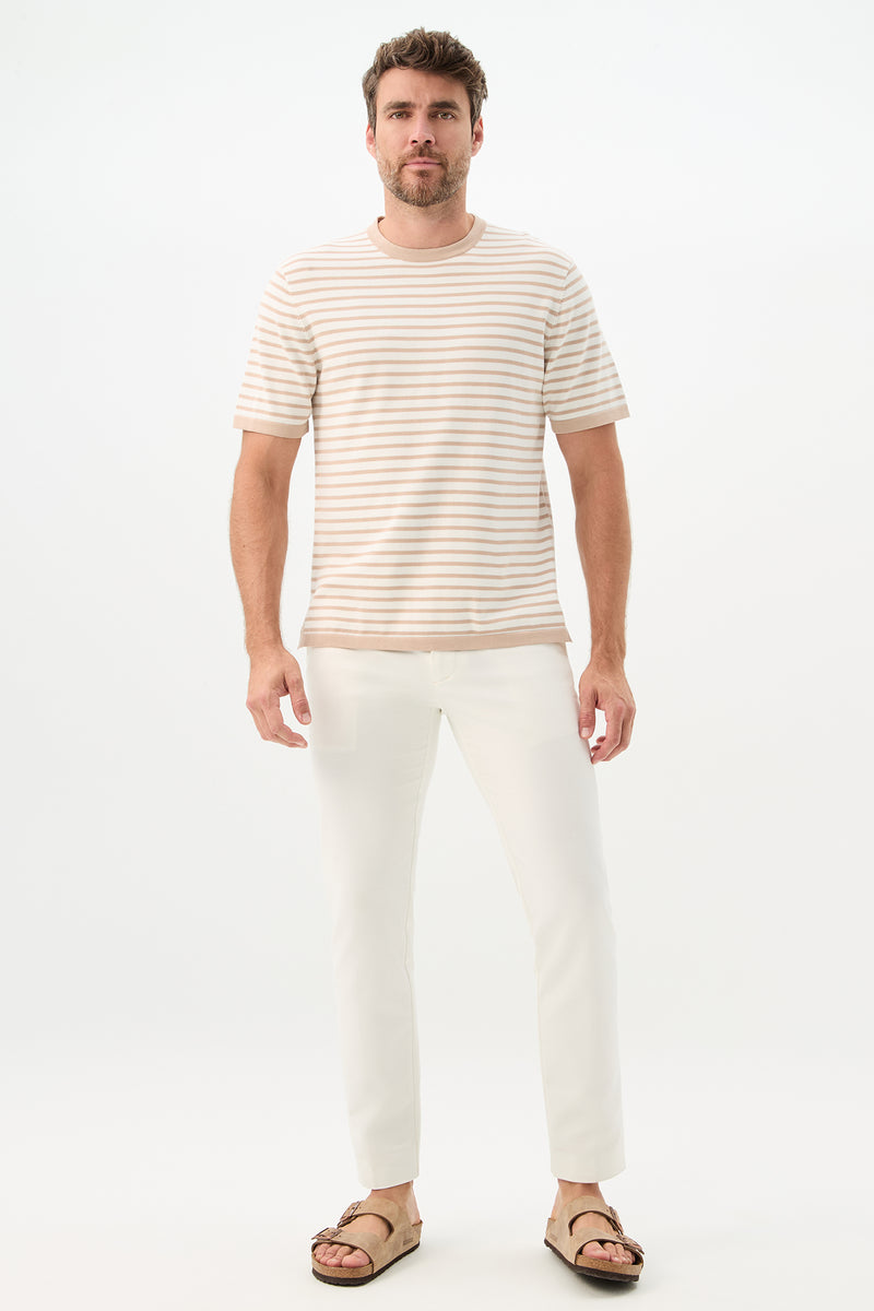 A man with short hair and a trimmed beard stands against a plain white background, wearing the Giovanni Stripe Short-Sleeve Pullover in beige and white stripes, paired with white pants and brown sandals, looking straight ahead with a neutral expression.