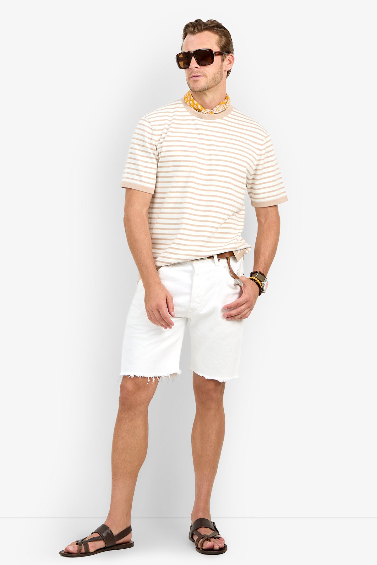 A man wearing sunglasses, the Giovanni Stripe Short-Sleeve Pullover, white frayed shorts, brown sandals, a watch, and a yellow scarf stands against a white background.
