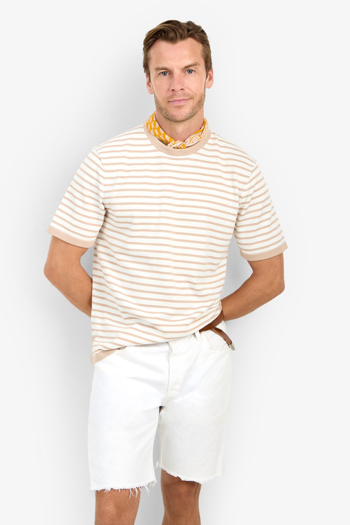 A man in the Giovanni Stripe Short-Sleeve Pullover, white frayed shorts, and a yellow patterned neck scarf stands against a plain white background with his hands behind his back and a slight smile.