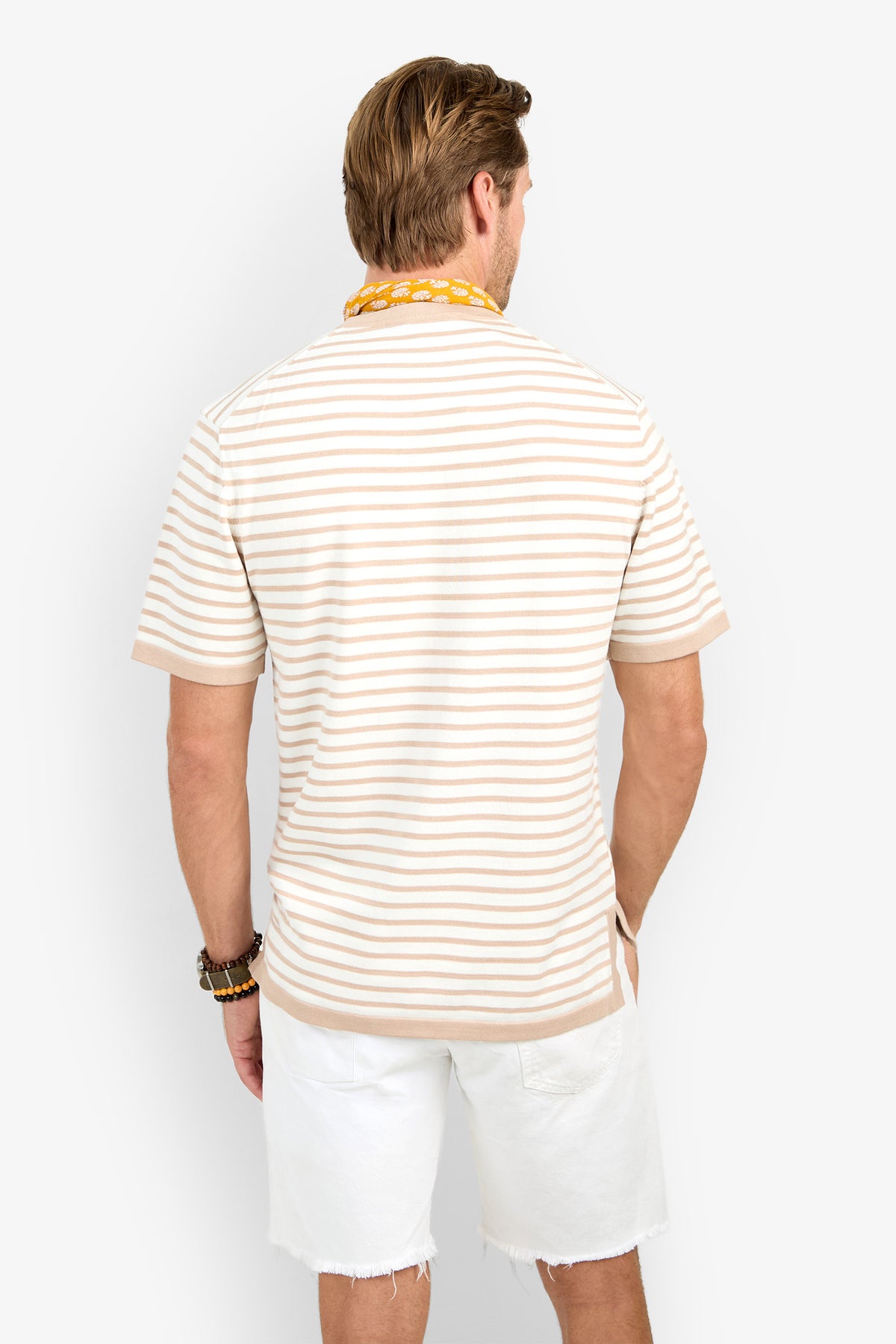 A man faces away, wearing the Giovanni Stripe Short-Sleeve Pullover in beige and white stripes, paired with white frayed shorts, bracelets, and a yellow beaded necklace against a plain white background.