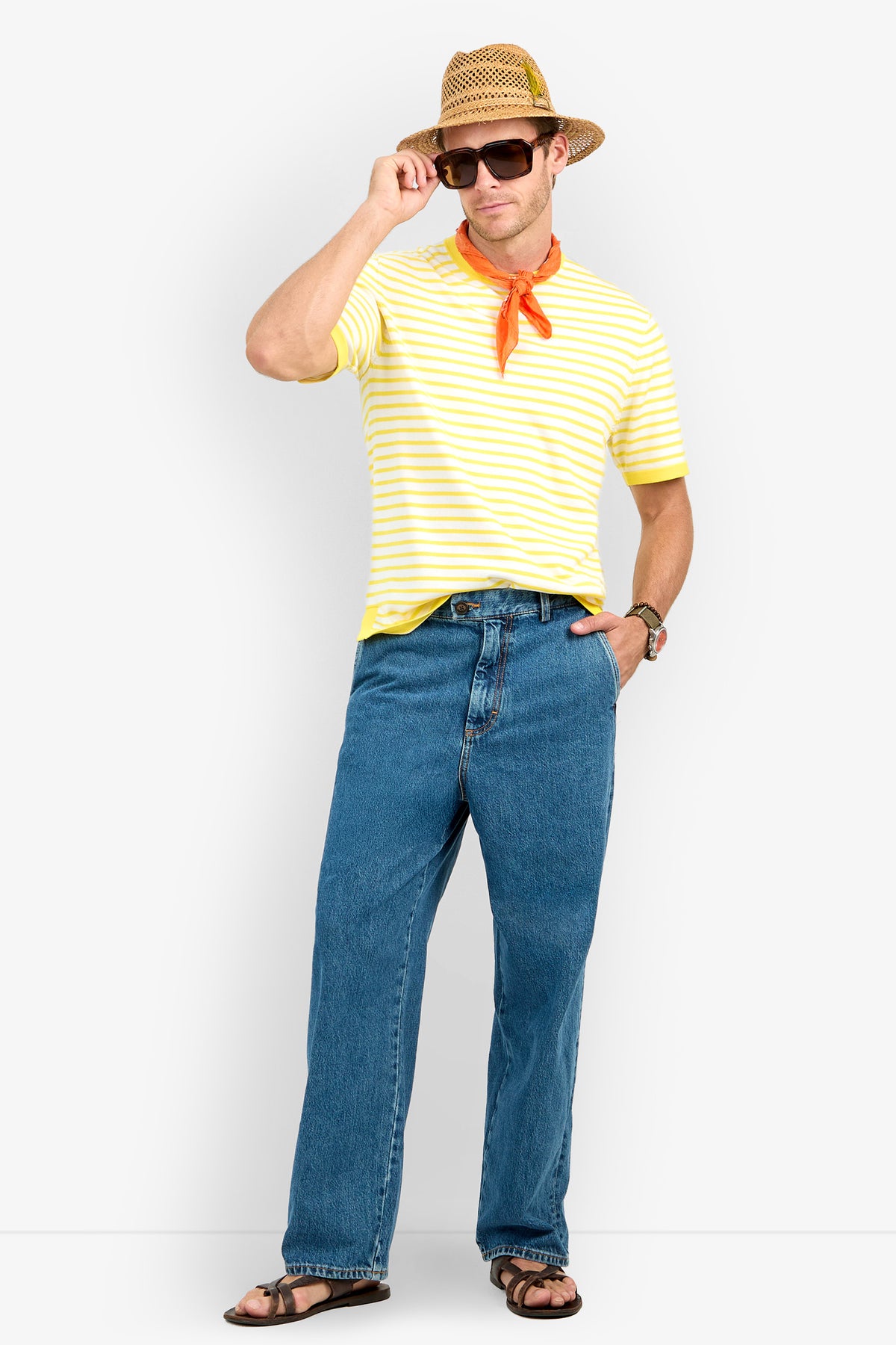A man in a straw hat, sunglasses, Giovanni Stripe Short-Sleeve Pullover, orange neck scarf, blue jeans, watch, and sandals stands against a white background, one hand adjusting his hat.