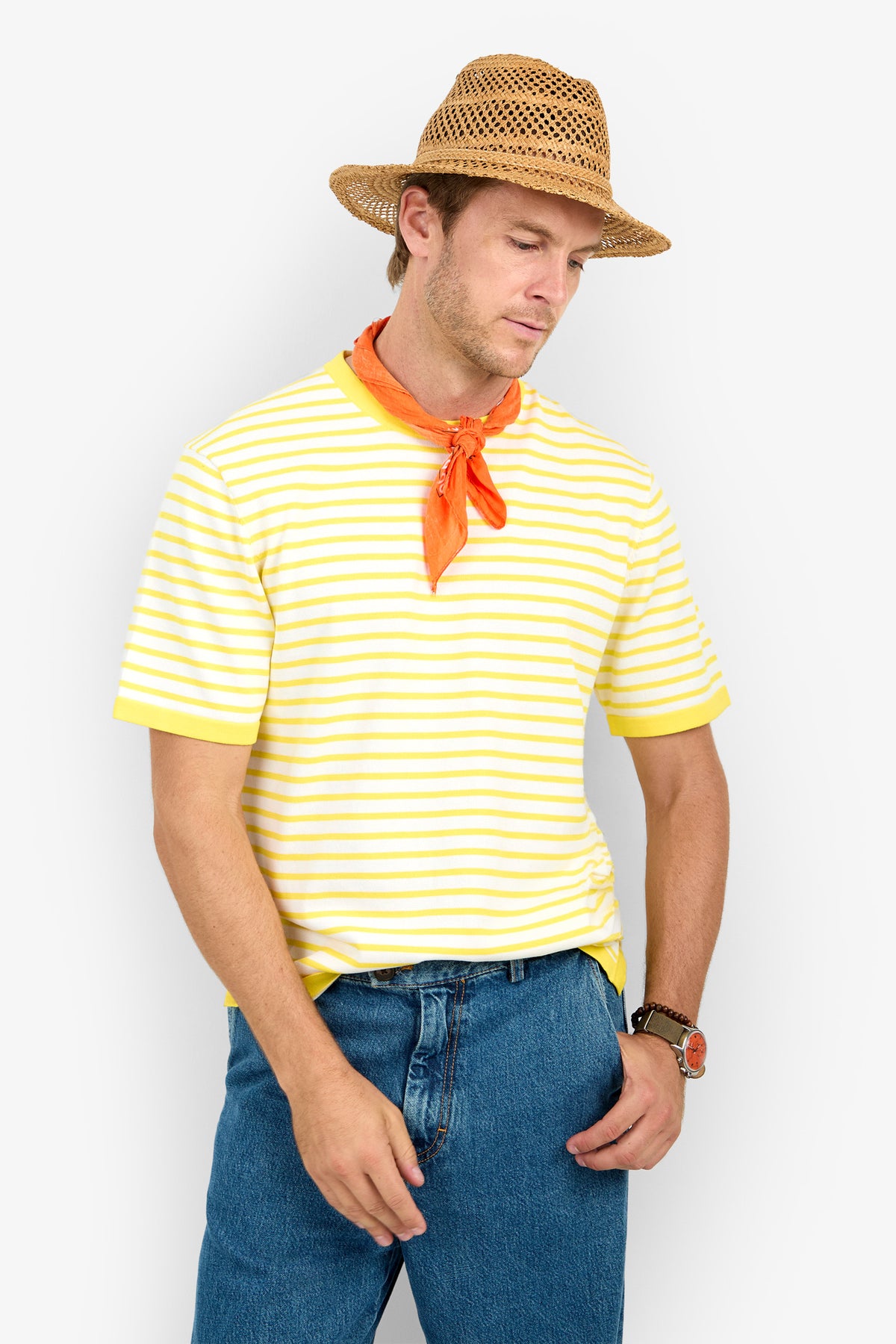 A man in a straw hat, orange neckerchief, blue jeans, and wristwatch wears the Giovanni Stripe Short-Sleeve Pullover, standing against a white background with one hand in his pocket, looking down.