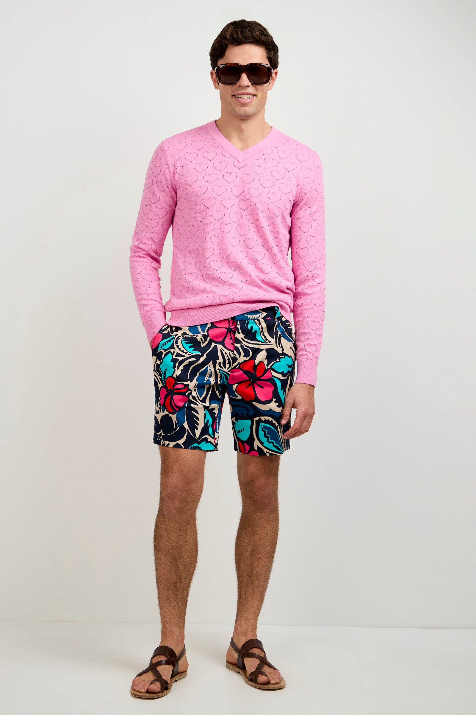 A man wears the Eros Heart Pointelle V-Neck Sweater, colorful floral shorts, brown sandals, and sunglasses, standing and smiling against a white background.