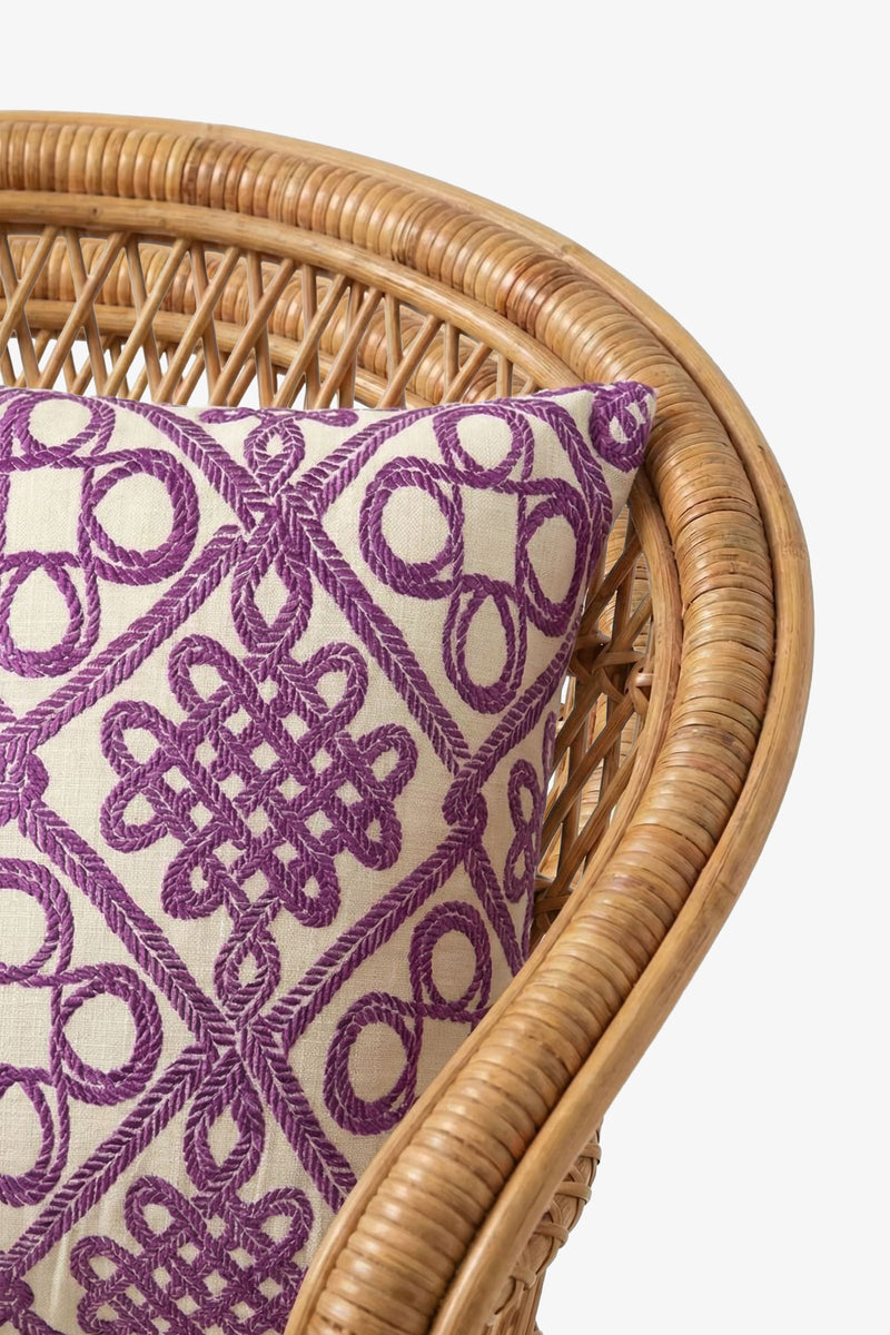 A close-up of a rattan chair with the Embroidered Square Pillow in beige and purple, featuring geometric and knot-like patterns.