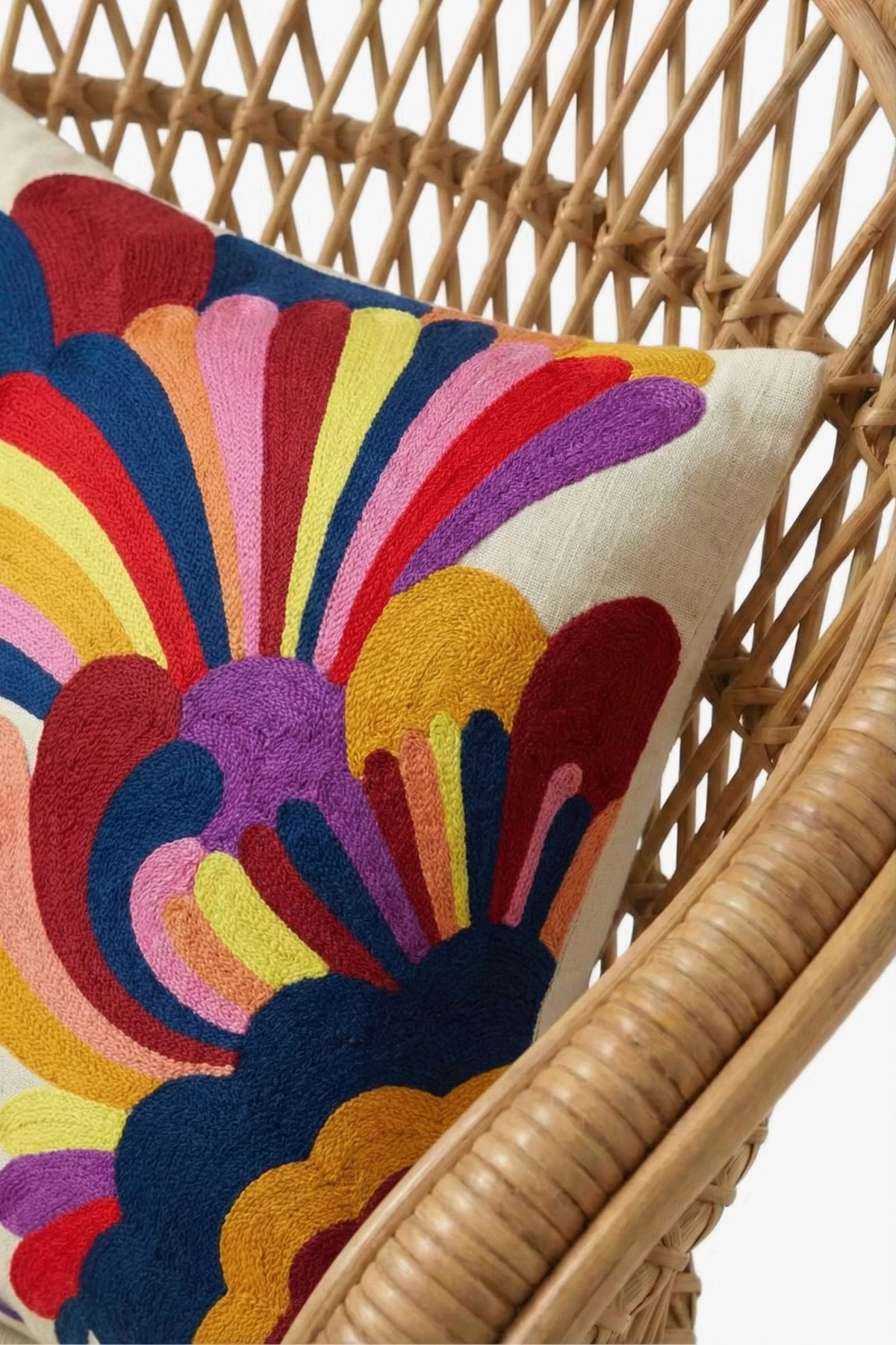 An Embroidered Lumbar Pillow, featuring a white base adorned with colorful, abstract floral patterns in red, yellow, blue, pink, purple, and orange, rests on a rattan chair.