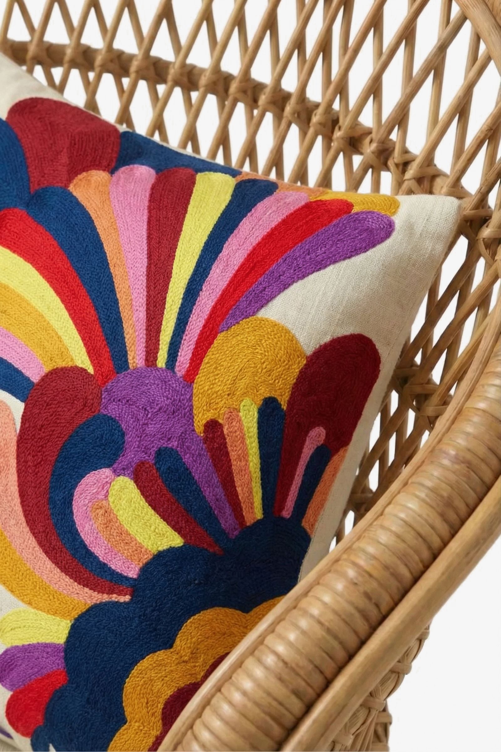 An Embroidered Lumbar Pillow, featuring a white base adorned with colorful, abstract floral patterns in red, yellow, blue, pink, purple, and orange, rests on a rattan chair.