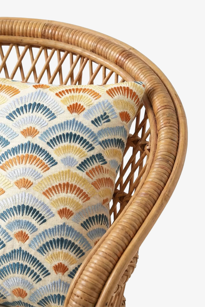A close-up of a wicker chair adorned with the Pasadena Embroidered Square Pillow, which features a decorative fan-patterned design in blue, orange, yellow, and gray tones.