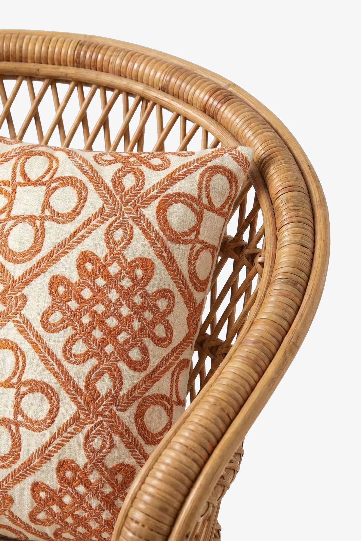 A close-up of a rattan chair with a beige cushion featuring the Costa Mesa Embroidered Square Pillow, adorned with an intricate, geometric orange embroidered pattern. The chair has a curved back with woven detailing.