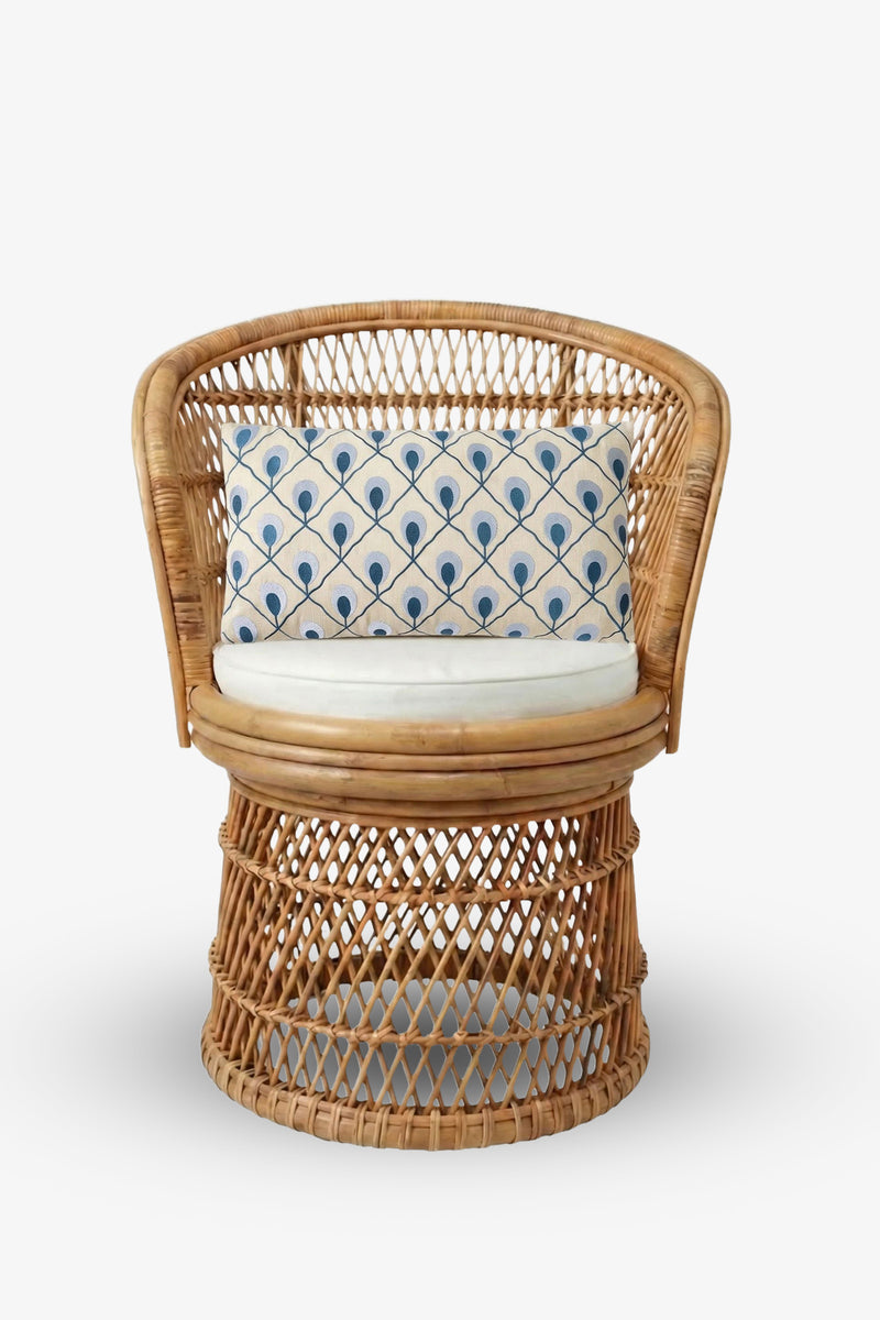 A round rattan chair with a woven design, a white seat cushion, and the Roseville Embroidered Lumbar Pillow featuring blue peacock feather patterns on a cream background.