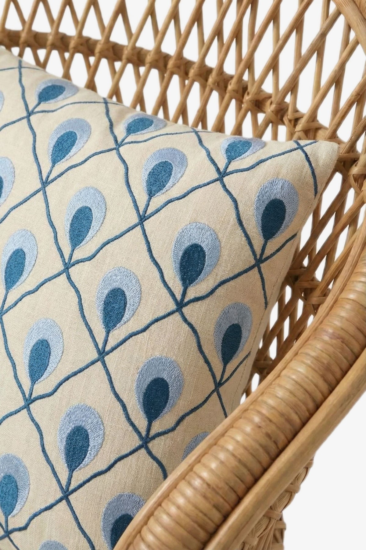 A close-up of a rattan chair with a cream cushion accented by the Roseville Embroidered Lumbar Pillow, featuring a blue peacock feather-inspired pattern.