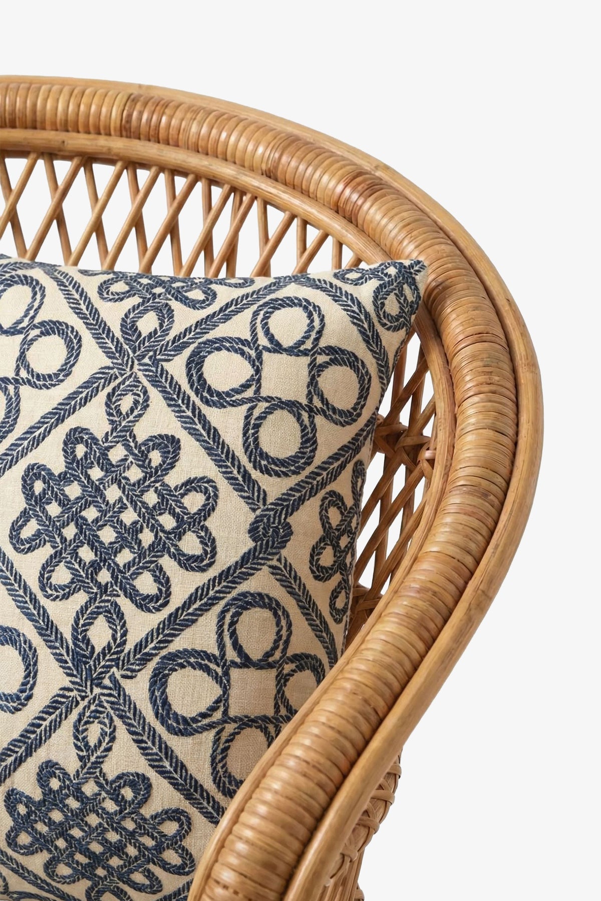 A close-up of a wicker chair with the Costa Mesa Embroidered Square Pillow, which features a blue geometric rope pattern on a cream fabric background.