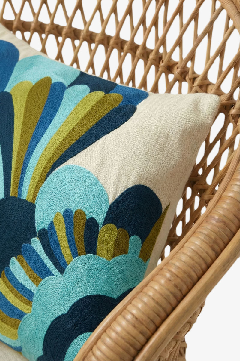 A close-up of a wicker chair with the Escondido Embroidered Lumbar Pillow, showcasing bold, embroidered blue, teal, olive, and navy abstract floral designs on a cream background.