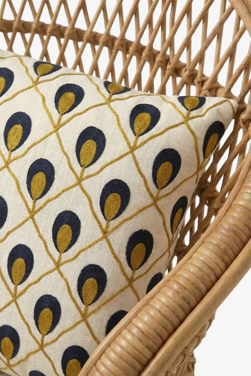 A close-up of the Roseville Embroidered Lumbar Pillow, featuring a gold and navy teardrop pattern, resting on a wicker chair.