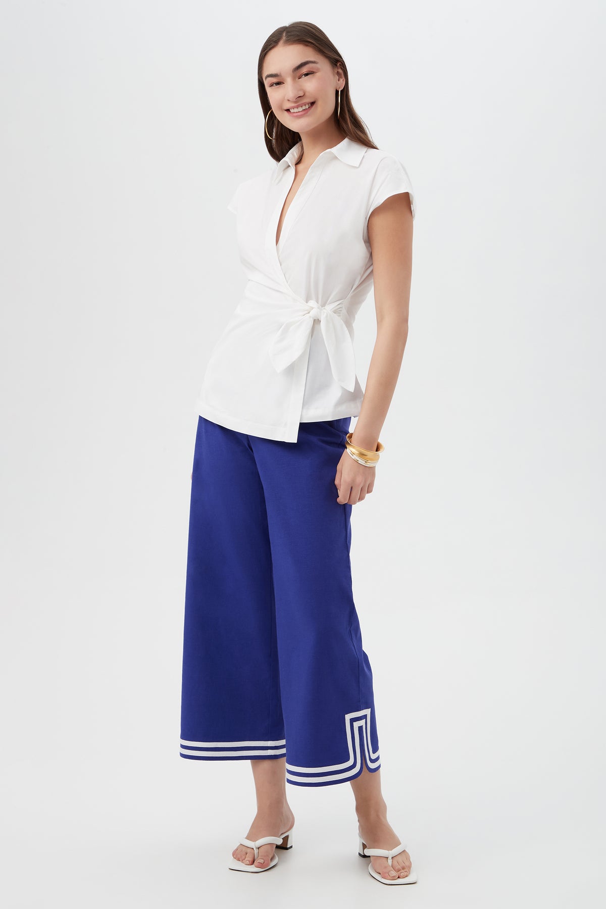 A woman stands smiling in the Avant Top, a white short-sleeve wrap blouse, paired with cobalt blue wide-leg culottes with white-trimmed hems, white sandals, and gold bracelets against a plain white background.