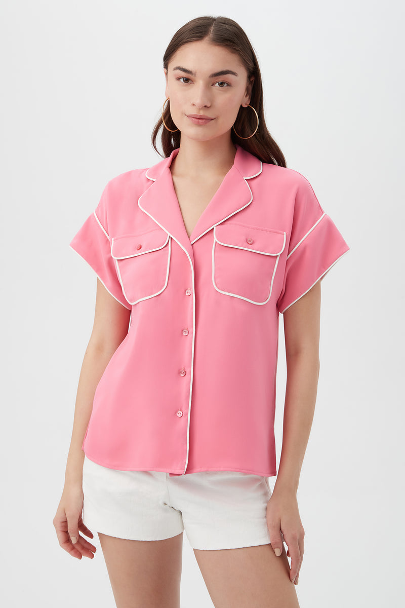 A woman wears the Gideon 2 Top in Eden Roc Rose, a short-sleeved button-up with white piping and two front pockets, paired with white shorts, against a plain light background.