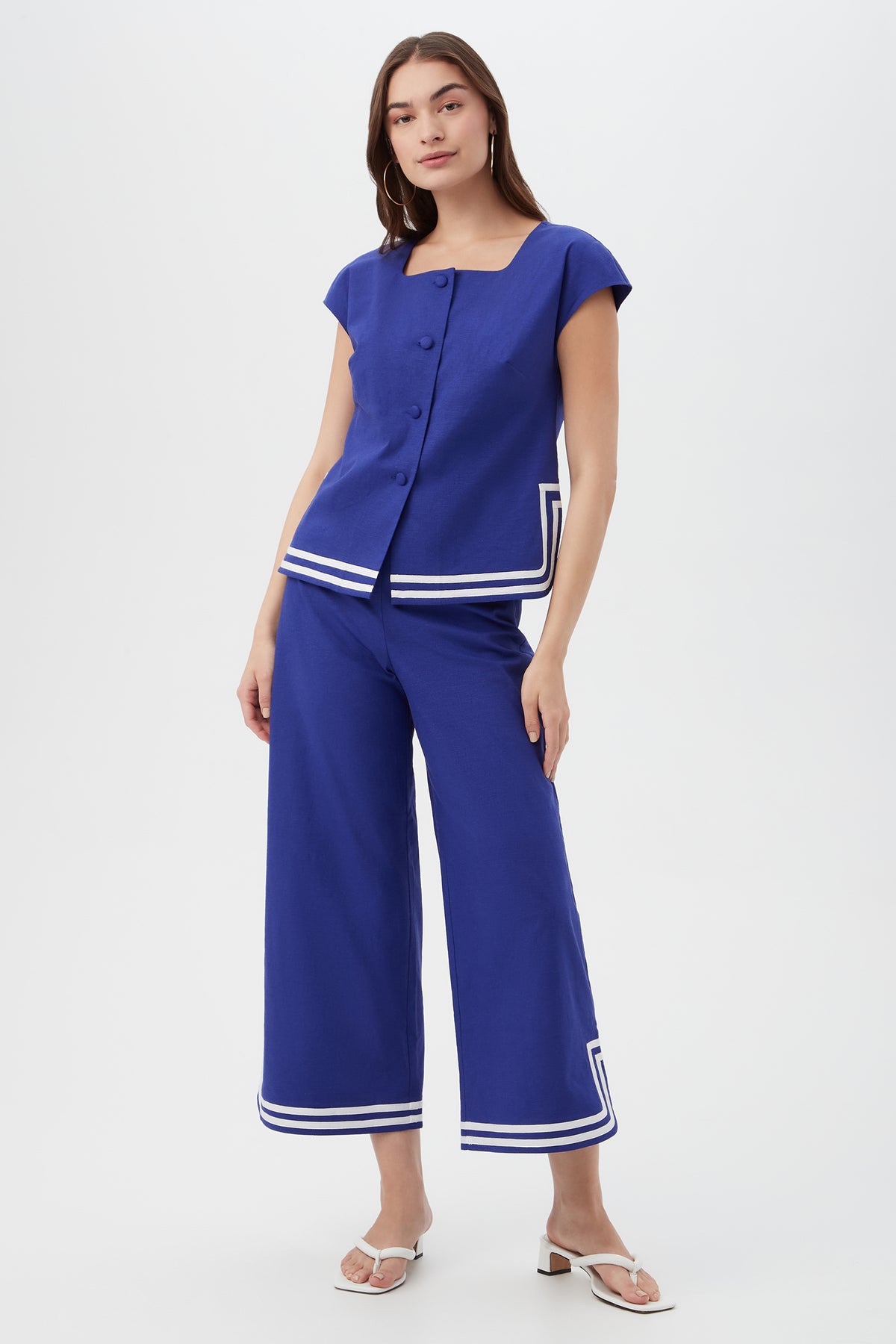 A woman wears the Valence Top, a blue short-sleeve shirt with large buttons and white stripe details near the hem, paired with matching wide-leg pants and white sandals, standing against a plain background.