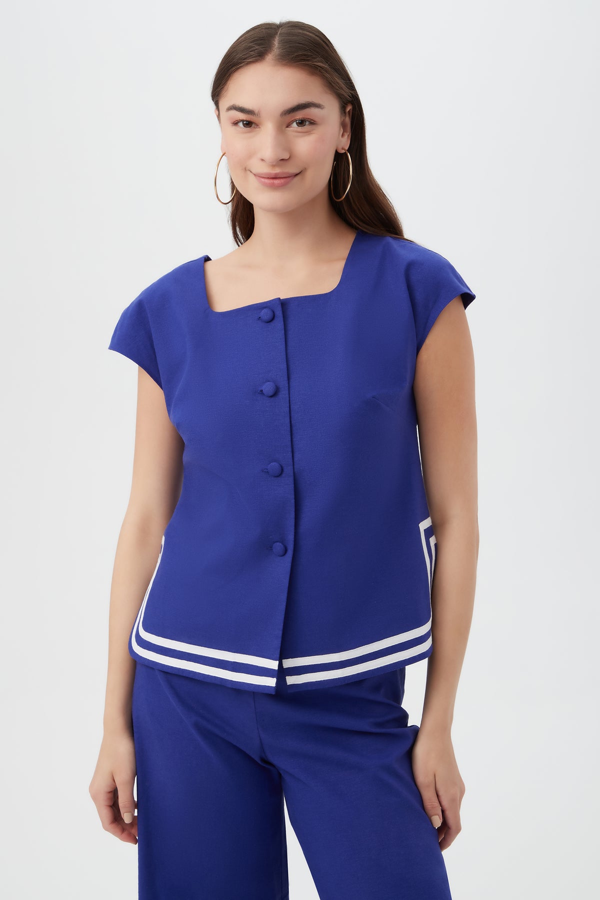 A woman wears the Valence Top in Cobalt Cove, featuring short sleeves, front buttons, and white stripe details at the hem, paired with matching blue pants. She has long dark hair, hoop earrings, and is smiling.