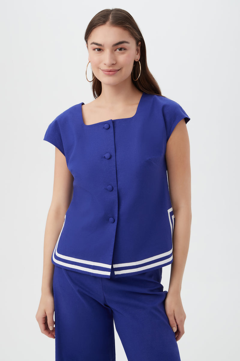 A woman wears the Valence Top in Cobalt Cove, featuring short sleeves, front buttons, and white stripe details at the hem, paired with matching blue pants. She has long dark hair, hoop earrings, and is smiling.