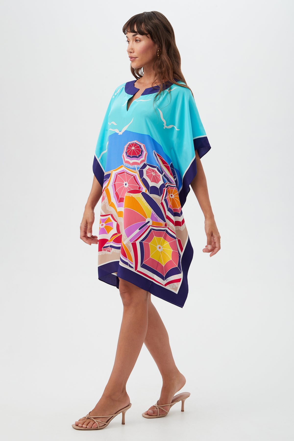 A woman models the Theodora Caftan Dress ‚Äö√Ñ√¨ Parapluie Silk, featuring vibrant umbrella and ocean motifs, paired with heeled sandals against a plain white background.