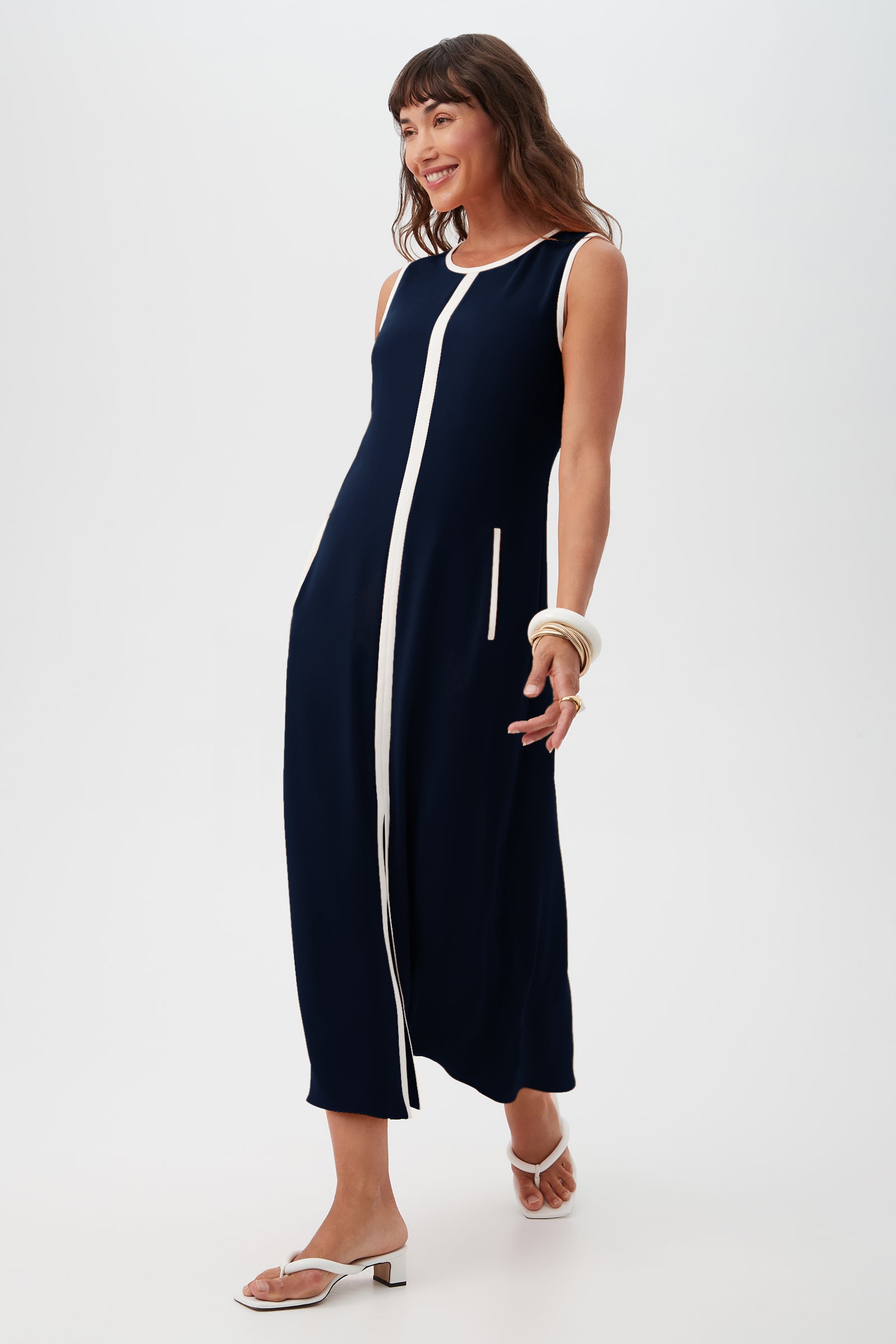 A woman wearing the Magnifique Dress—a sleeveless navy blue midi with a white vertical front stripe—paired with white sandals and bangles, smiles while walking against a plain white background.