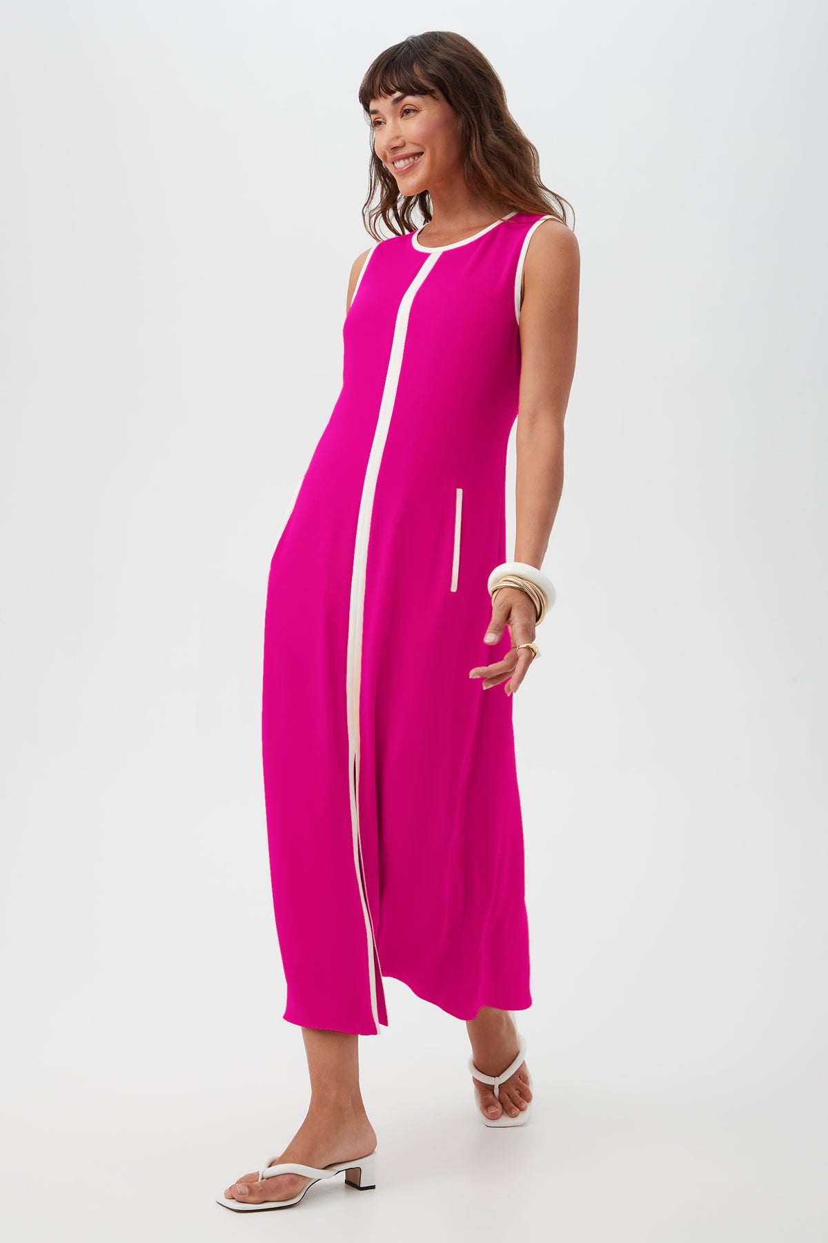 A woman with long brown hair smiles and walks against a white background, wearing the Magnifique Dress—a sleeveless bright pink midi dress with a white vertical stripe—white sandals, and white bangles.