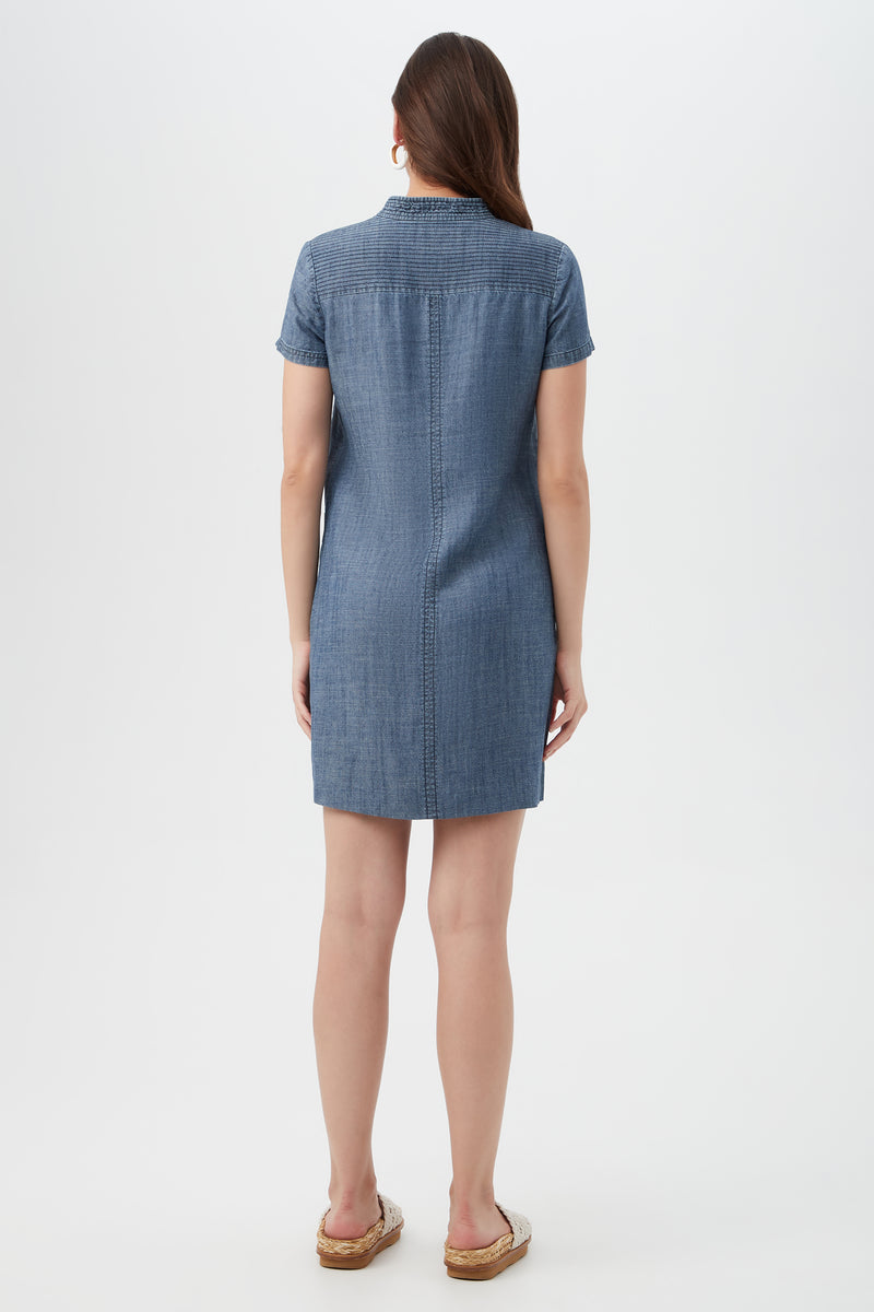 A woman faces away from the camera in a Suntan 2 Short-Sleeve Chambray Dress and brown sandals, with a plain white background.