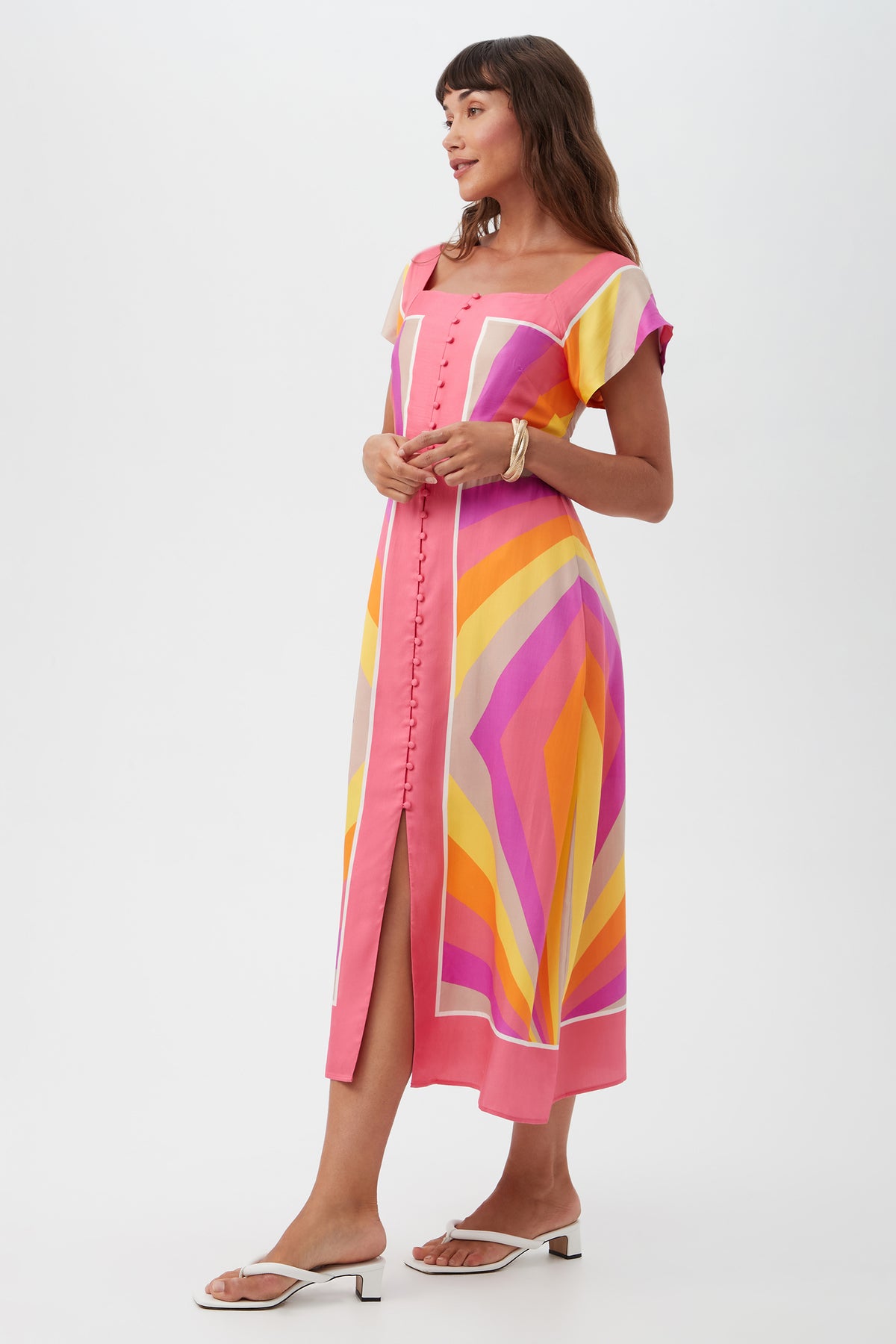 A woman stands sideways in the Après Short-Sleeve Dress, a colorful midi with geometric patterns in pink, orange, yellow, and purple. It features short sleeves, front buttons, a slit, and she pairs it with white sandals and bracelets.