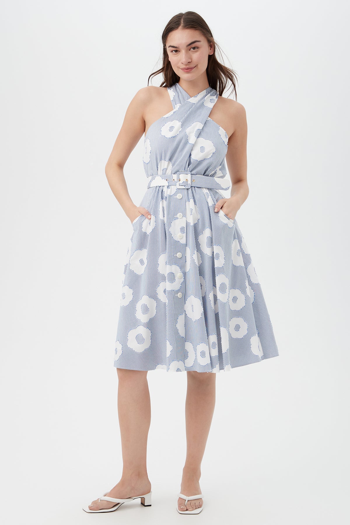 A woman wears the Mardi Sleeveless Dress in Cassis Cotton—a knee-length, belted dress with a halter neckline, pockets, and white floral patterns—smiling with hands in pockets and paired with white sandals.