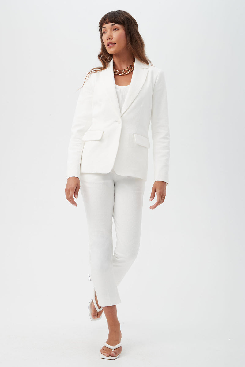 A woman with long brown hair stands against a white background, wearing Pop Slim-Leg Stretch Pants, a white blazer, white top, white sandals, and a chunky necklace. She looks slightly to the side.