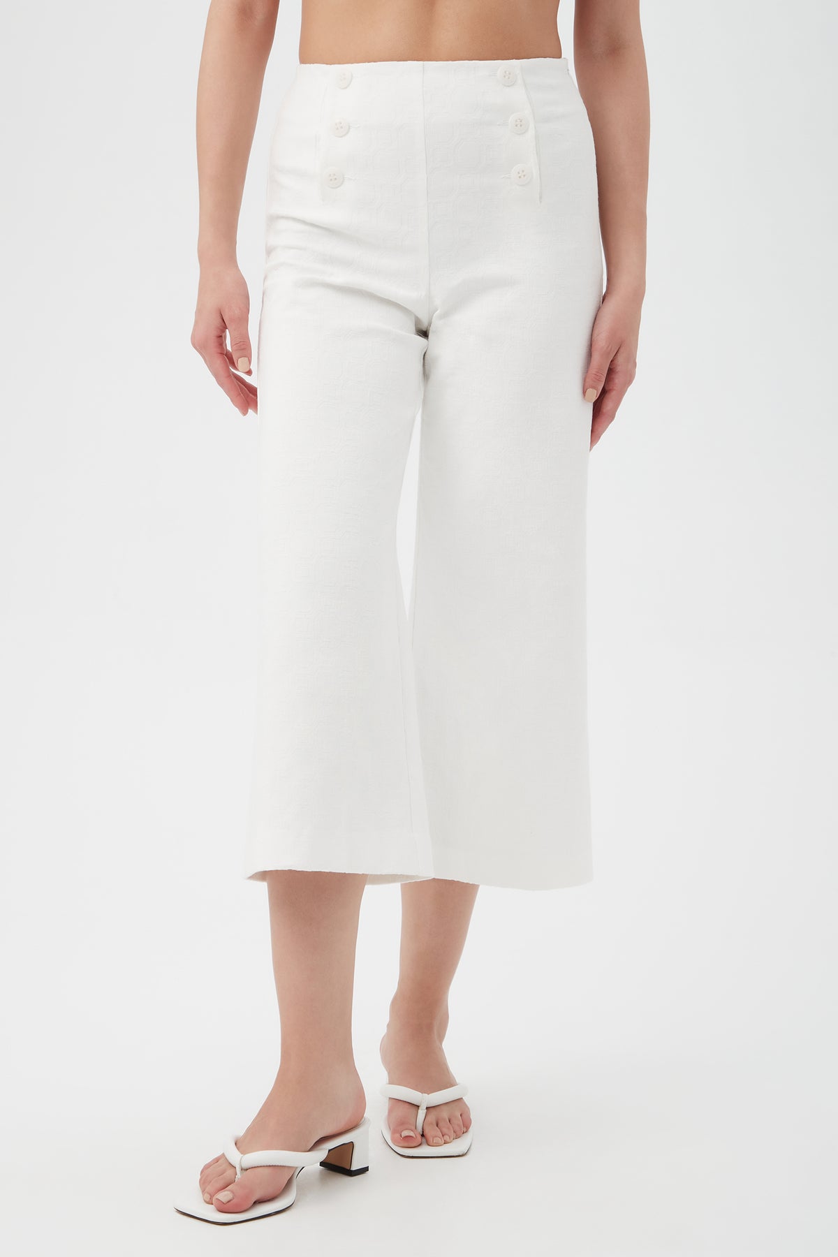 Someone wearing Saltie 2 Crop Pants in Whitewash with button details and white heeled sandals stands against a plain white background. Only their lower torso and arms, relaxed at their sides, are visible.
