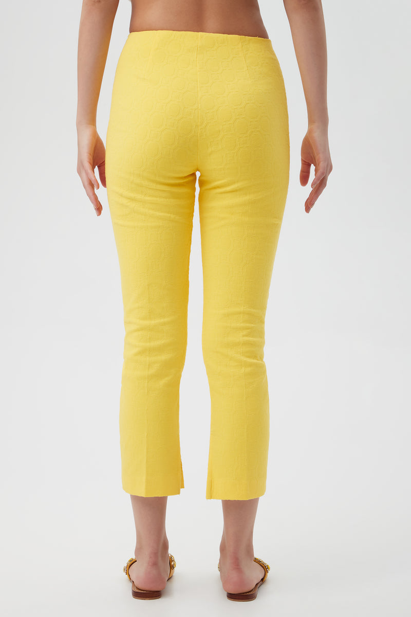 A person faces away from the camera, wearing Pop Slim-Leg Stretch Pants in bright yellow with a textured finish, cropped hem, and back slits, styled with flat sandals against a plain white background.