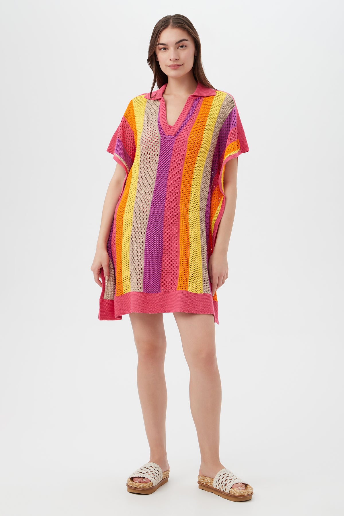 A woman with long brown hair wears the Candela Dress in Eden Roc Rose—a loose, short-sleeve crochet style with vertical stripes—paired with white sandals, standing against a plain white background.