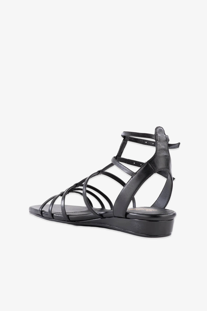 The Luxurious Wedge Sandal in black leather features a strappy flat design with a slight heel, an open toe, and multiple thin straps crossing over the foot and wrapping around the ankle, shown against a white background.