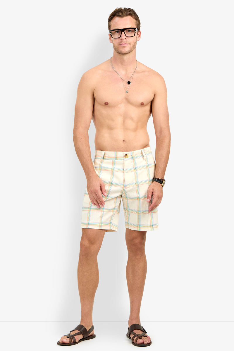 A man in glasses, a necklace, a watch, sandals, and the Travis Short - Pride Plaid stands against a plain background with his arms relaxed at his sides.