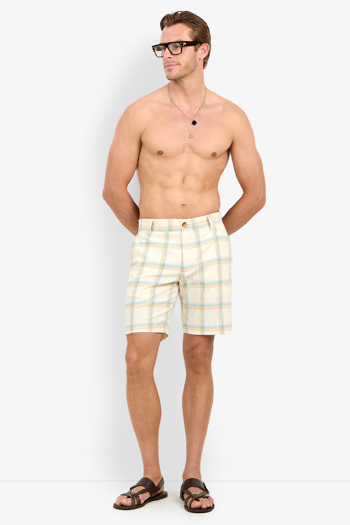 A shirtless man in glasses, sandals, and Travis Short - Pride Plaid stands with hands in pockets against a plain white background.
