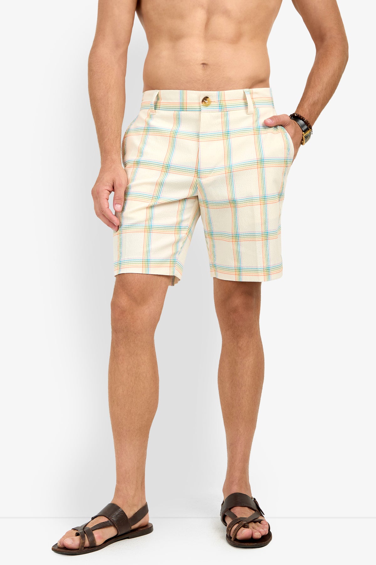 A man stands shirtless against a white background, wearing the Travis Short - Pride Plaid, a wristwatch, and brown sandals.
