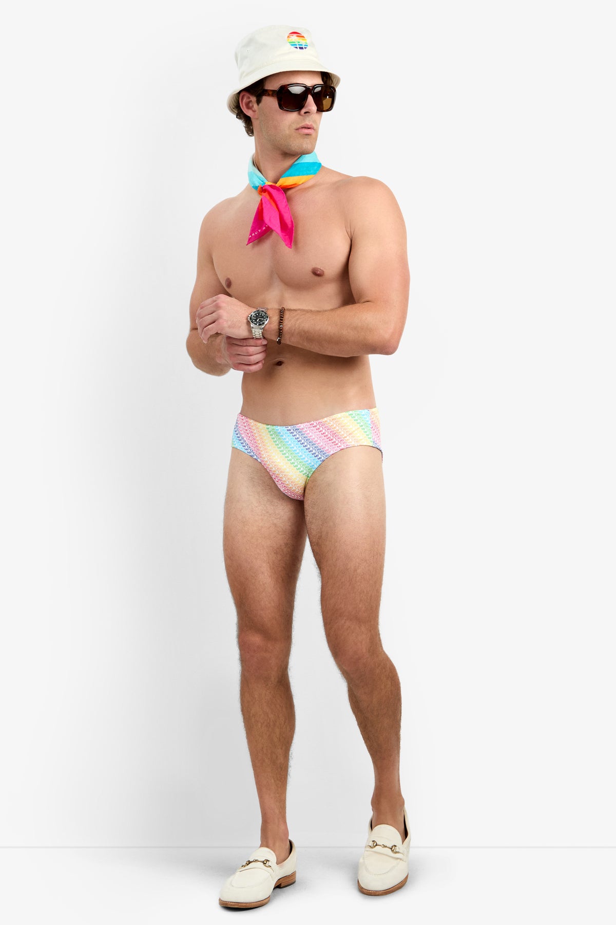 A man wearing sunglasses, a white bucket hat, a colorful neck scarf, the Bondi Swim Brief - Pride in a pastel rainbow pattern, and white loafers stands against a plain white background.
