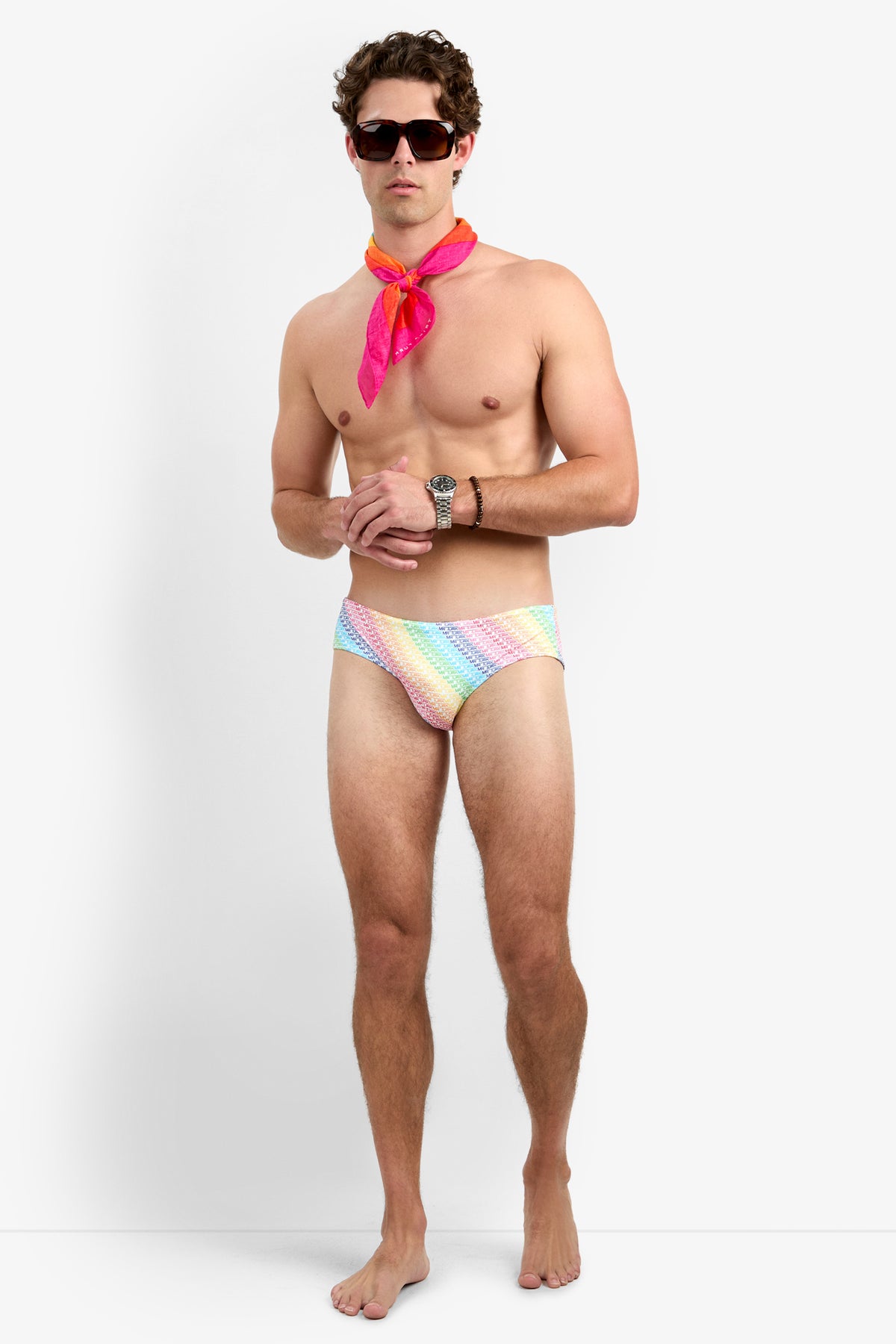 A man with short, curly brown hair and a neutral expression stands barefoot against a white background, wearing sunglasses, a pink scarf, a wristwatch, and the Bondi Swim Brief - Pride featuring a vibrant rainbow design.