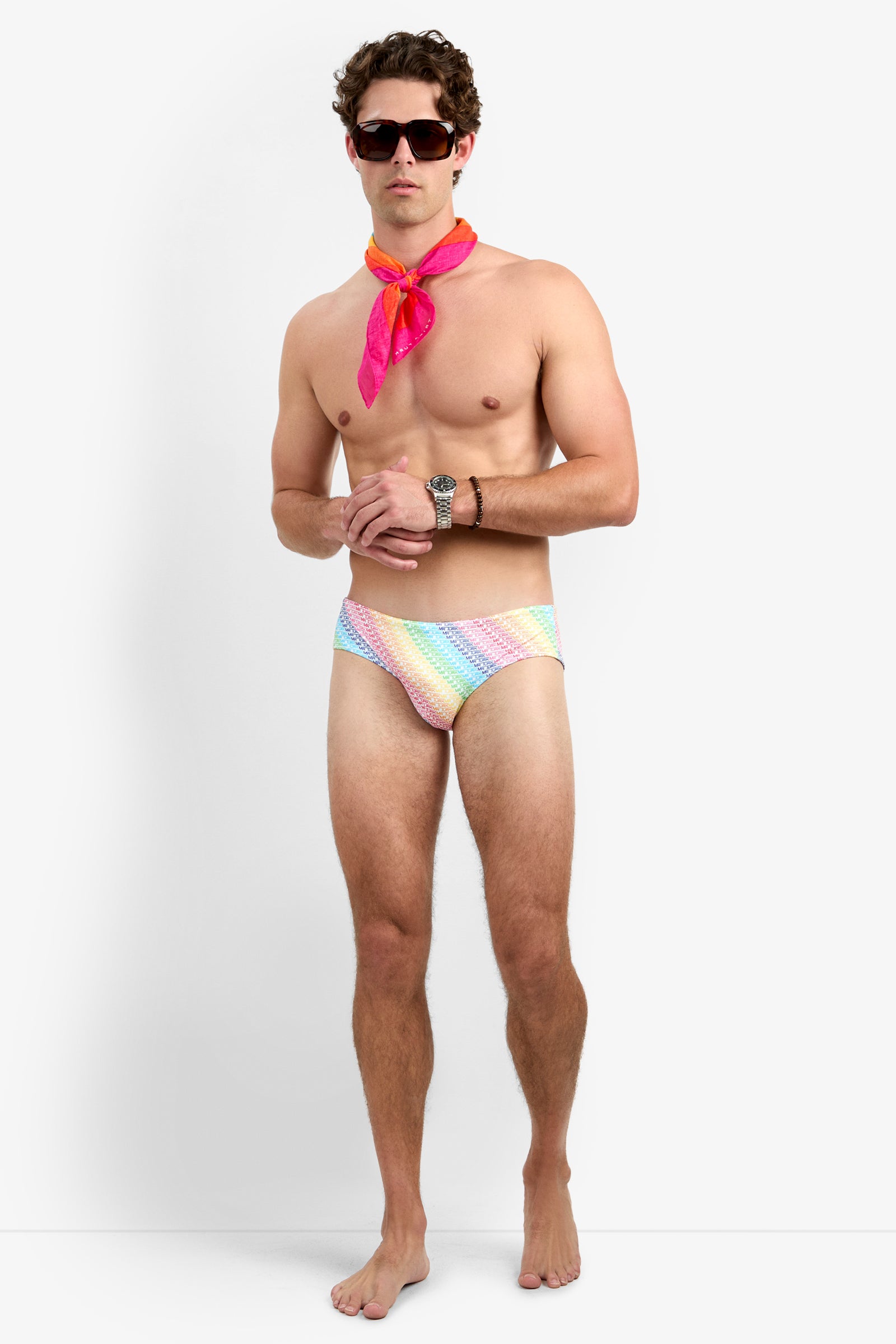 A man with short, curly brown hair and a neutral expression stands barefoot against a white background, wearing sunglasses, a pink scarf, a wristwatch, and the Bondi Swim Brief - Pride featuring a vibrant rainbow design.