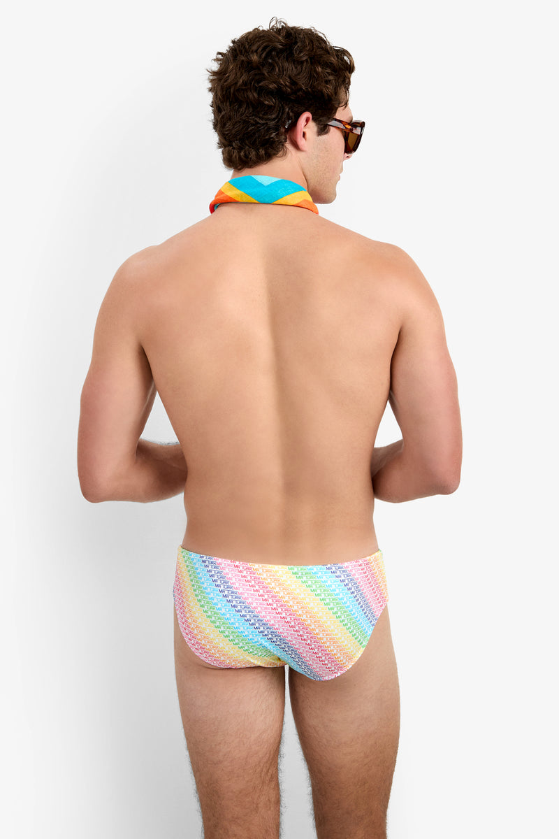A man with curly brown hair faces away from the camera, wearing sunglasses, a rainbow-striped neck scarf, and the Bondi Swim Brief - Pride against a plain white background.