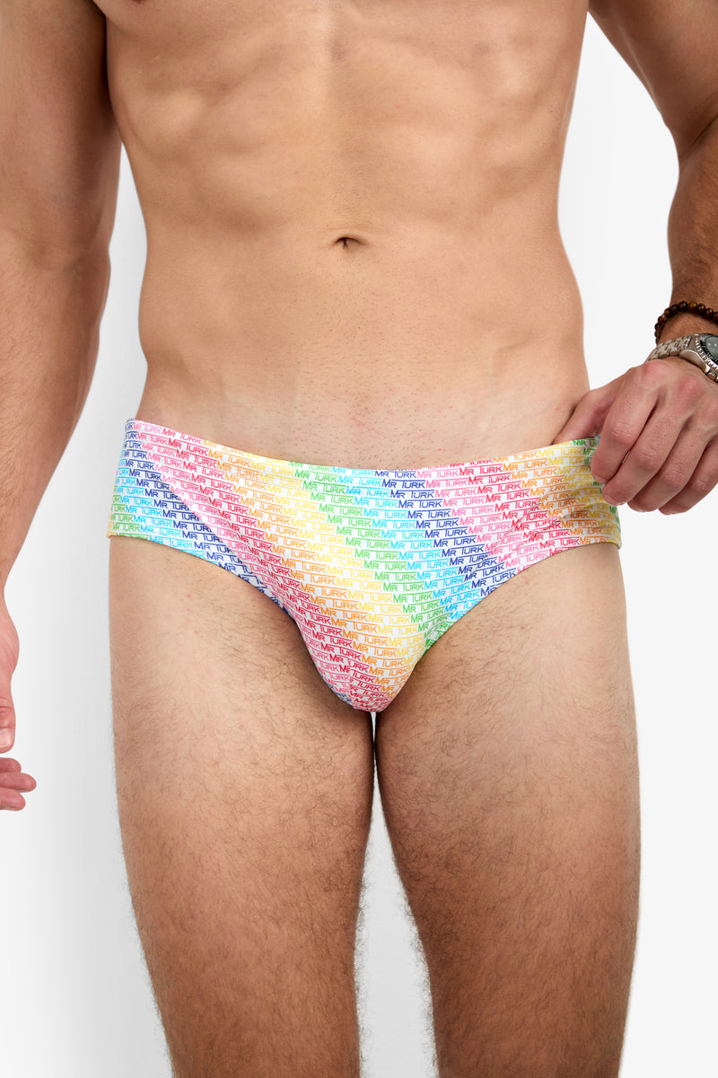 A man models the Bondi Swim Brief - Pride, featuring a rainbow gradient with repeating white text. His torso and upper thighs are visible as he holds the waistband with one hand.
