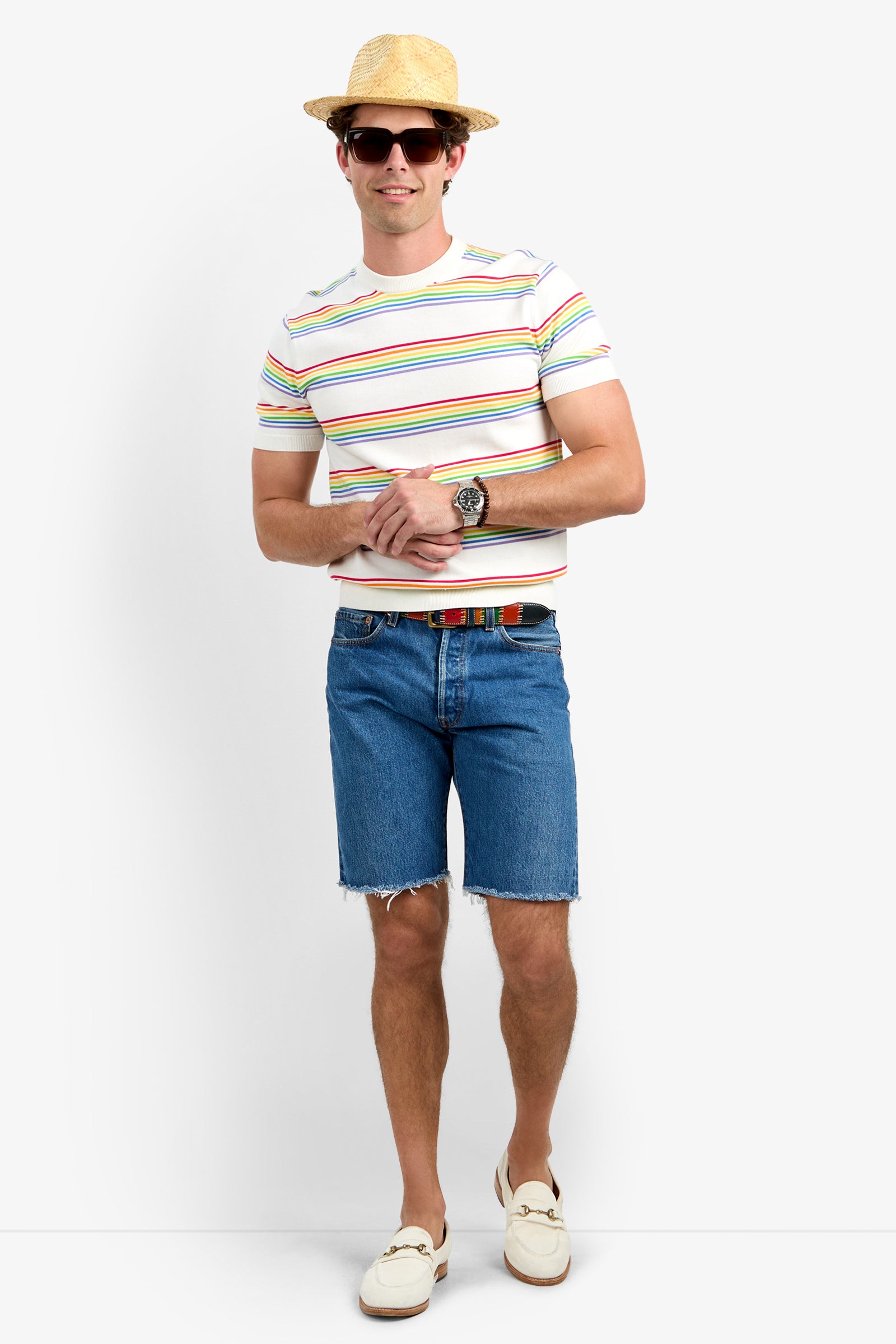 Wearing the Carlo Short-Sleeve Crewneck - Pride, a smiling person in a straw hat, dark sunglasses, blue denim shorts, and white loafers stands with hands clasped in front.