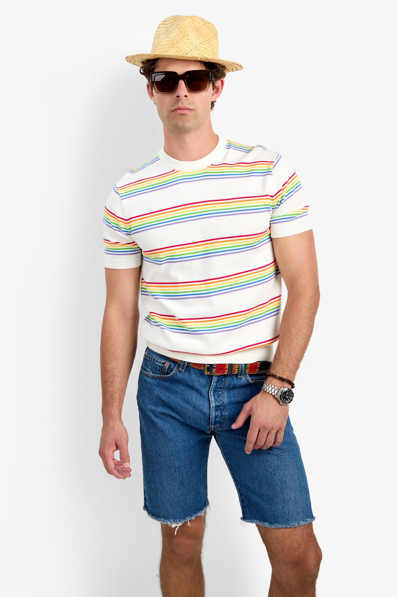 Man wearing the Carlo Short-Sleeve Crewneck - Pride, a straw hat, sunglasses, denim shorts, a multicolored belt, and a wristwatch stands against a plain white background.