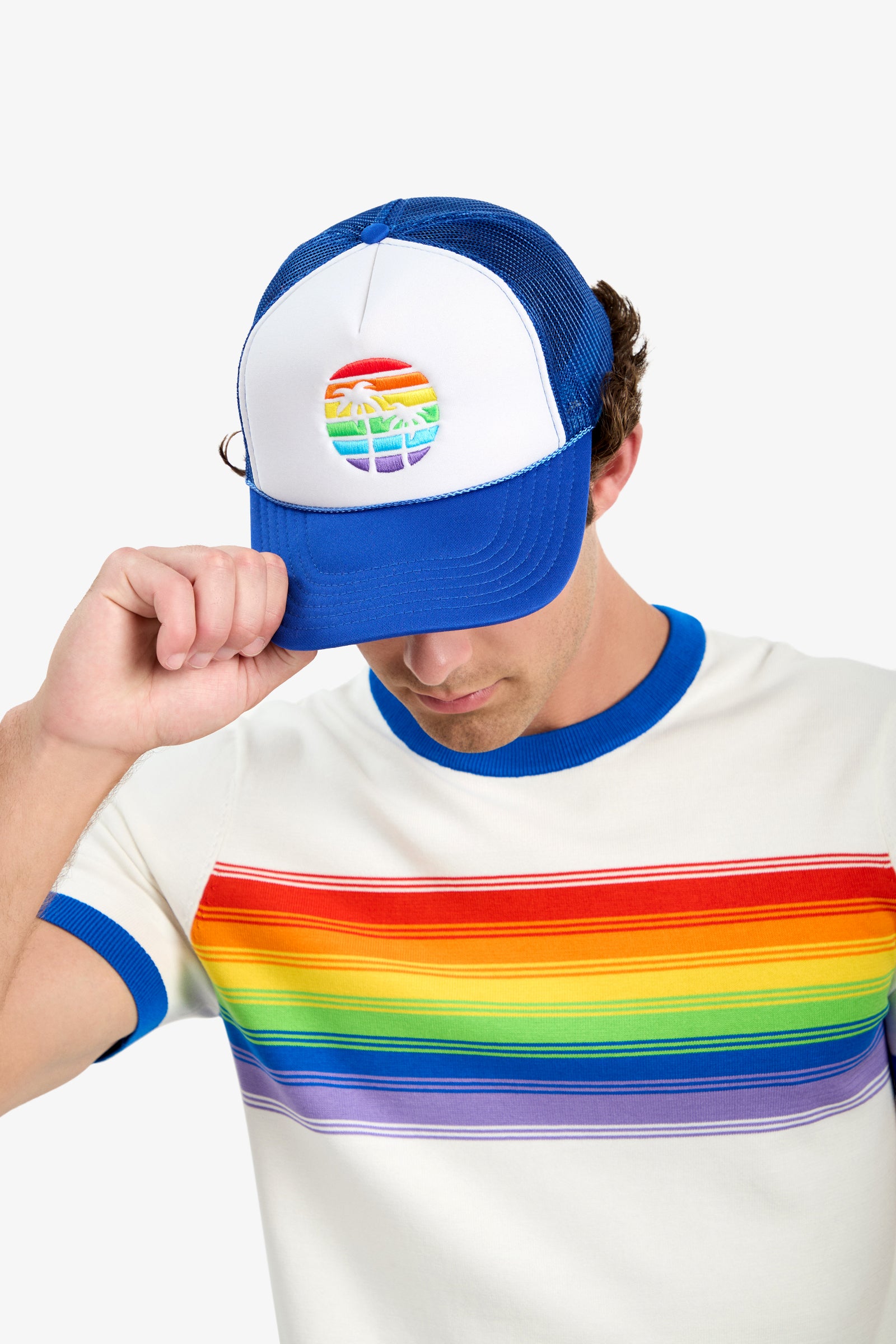 A person in a white T-shirt with a rainbow stripe and wearing the Trucker Cap - Pride, which features a rainbow and palm tree graphic, tilts the cap down with one hand.