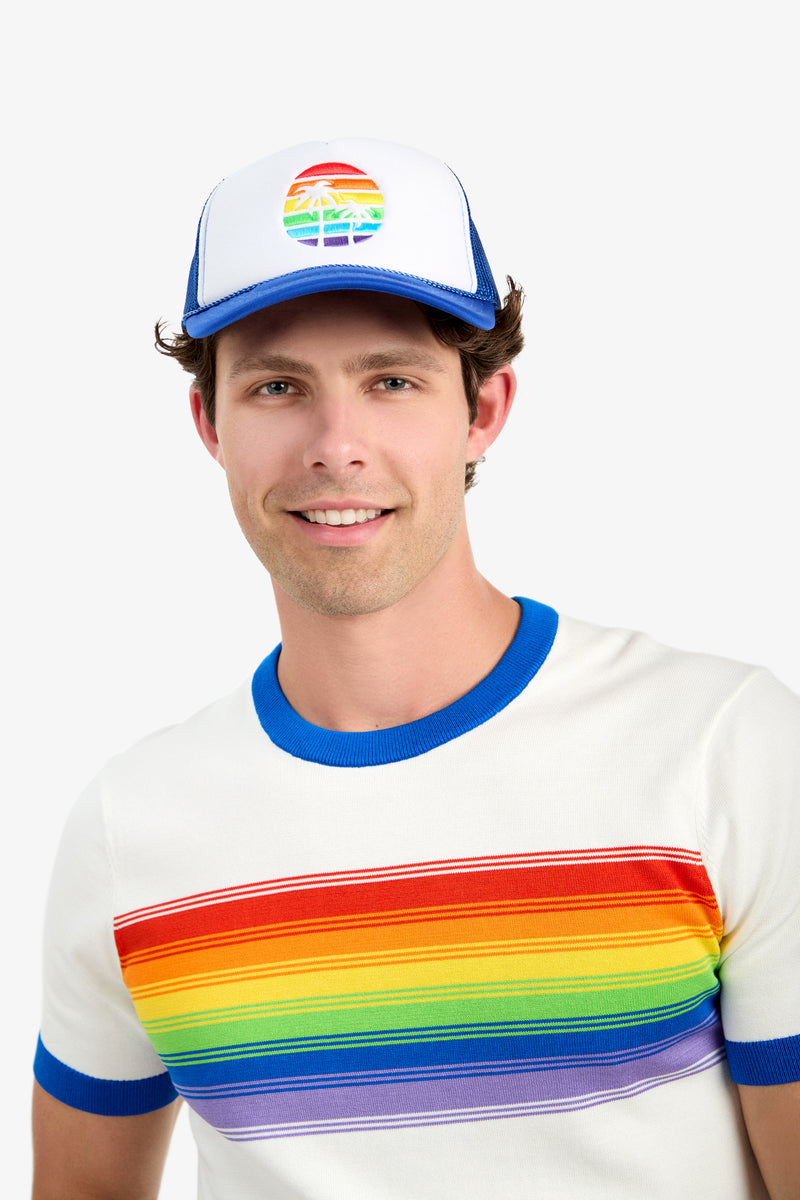 A smiling man wears the Trucker Cap - Pride, a white and blue hat with a rainbow and palm tree design, plus a white tee that has a bold horizontal rainbow stripe across the chest.