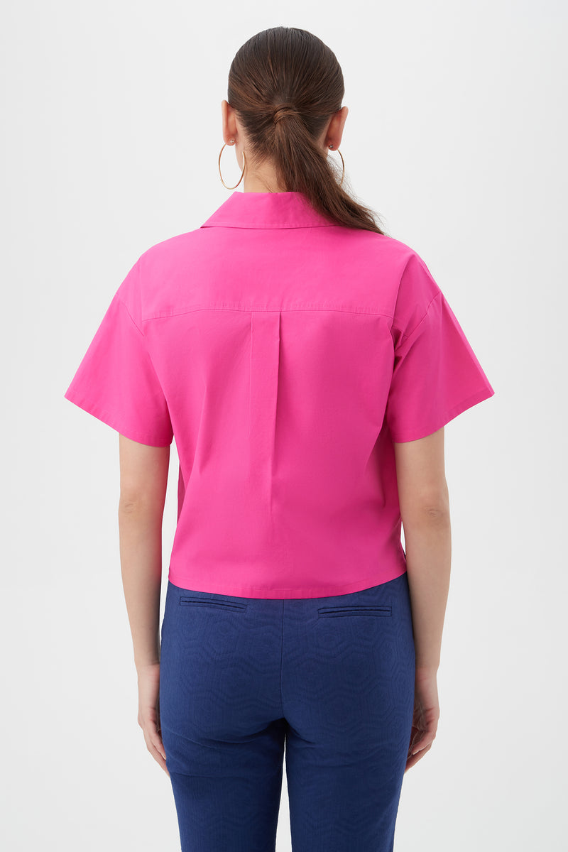 A woman is seen from behind in a Komore Cropped Button-Front Top‚Äö√Ñ√Æbright pink, short-sleeve‚Äö√Ñ√Æpaired with blue pants. She has straight brown hair tied back, wears large hoop earrings, and stands against a plain white background.