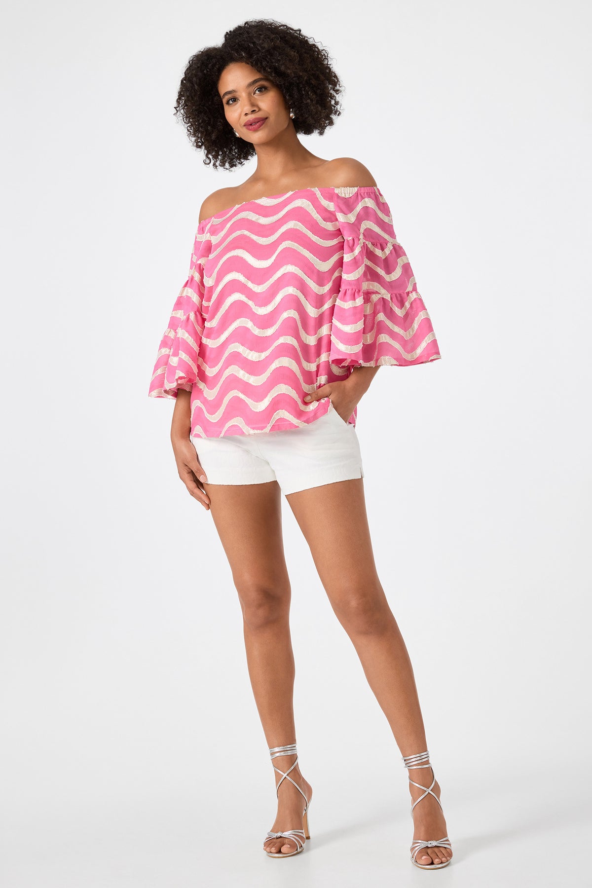 A woman stands against a plain white background wearing the Garnish 2 Off-Shoulder Top - Pinku, featuring pink and white wavy stripes and flared sleeves, paired with white shorts and strappy silver sandals.
